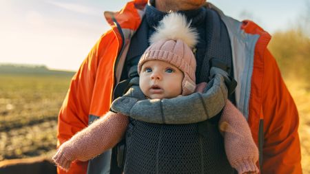A baby girl is shown being carried by her father in a baby carrier while out on a walk in the countryside. 